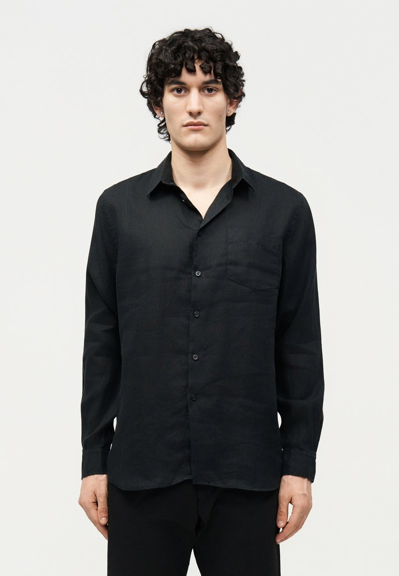 Νέος άνδρας με σγουρά σκούρα μαλλιά, που φοράει μακρυμάνικο μαύρο πουκάμισο με κουμπιά και μαύρο παντελόνι, μπροστά σε απλό λευκό φόντο.