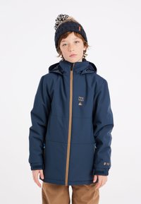 Veste imperméable bleue avec fermeture éclair à l'avant, accents beige, détail de logo et capuche ajustable. Portée avec un pantalon beige et un bonnet en tricot.
