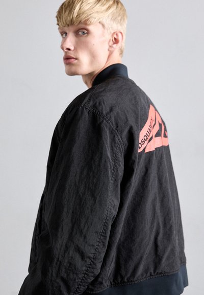 Blonde man die zijn hoofd draait, gekleed in een zwarte jas met een rood driehoekig logo en tekst op de rug, tegen een effen grijze achtergrond.