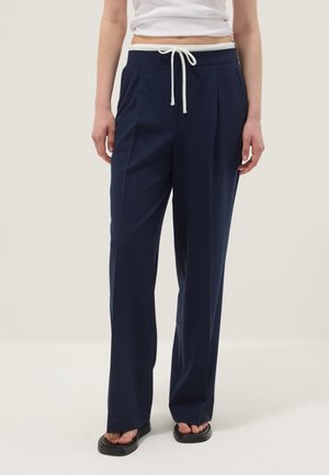 Persoon die marineblauwe wijde broek met wit trekkoord en zwarte slippers draagt, staand tegen een effen lichte achtergrond.