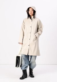 Beige vattentålig kappa med huva, knappstängning och sidofickor. Matchad med baggy jeans och svarta snörkängor. Texturerad accessoar inkluderad.