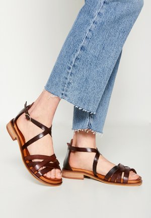 Füße, die braune flache Ledersandalen mit Riemchen tragen, kombiniert mit hellblauen Jeans mit ausgefranstem Saum vor neutralem Hintergrund.
