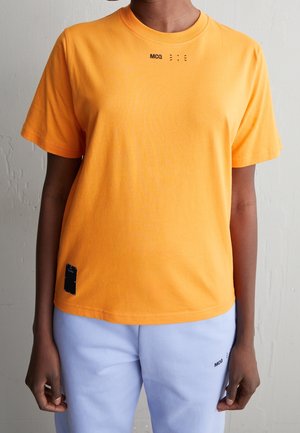 Osoba nosząca jaskrawo pomarańczowy t-shirt z krótkim rękawem z napisem "MCQ" na piersi oraz jasnoniebieskie spodnie z napisem "MCQ" w pobliżu kieszeni.