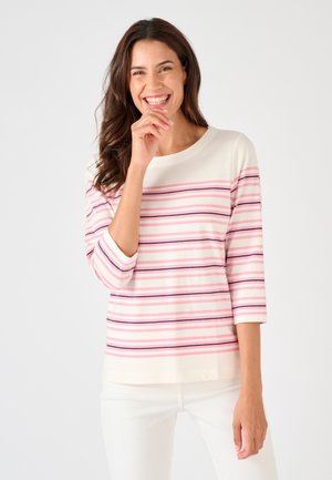 Lächelnde Frau mit langen braunen Haaren trägt ein weißes Oberteil mit pinken und marineblauen horizontalen Streifen und weiße Hose, steht vor einem weißen Hintergrund.