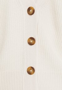 Krämfärgad stickad cardigan med djup V-ringning, fyra stora bruna marmorerade knappar, ribbad kant och strukturert tyg.