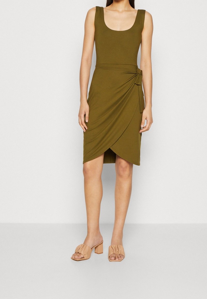 Robe longueur genoux vert olive avec un corsage ajusté et un design portefeuille, comprenant un détail de nœud sur le côté. Associée à des sandales à talons blocs beige clair.