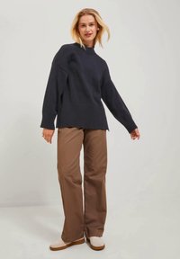 Suéter de punto azul marino con un cuello alto y dobladillo acanalado, combinado con pantalones de pierna ancha marrones y zapatos de color claro, de pie sobre un fondo neutro.