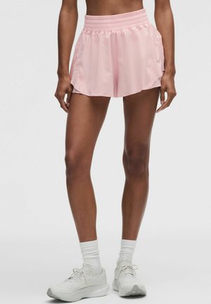Femme portant un short de sport rose clair, des chaussettes blanches et des chaussures de course blanches, debout devant un fond gris uni.