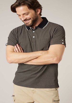 Homme les bras croisés portant un polo à manches courtes noir à motifs avec un col bordé de blanc et un pantalon cargo beige.