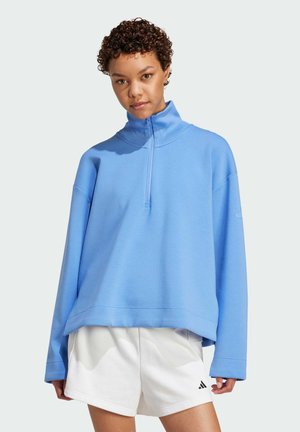 Felpa azzurra chiara con colletto alto, maniche lunghe e zip a metà. Indossata sopra pantaloni sportivi bianchi.