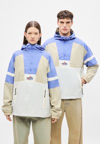 Kék, krémszínű és fehér színblokkos széldzseki félcipzáros elülső résszel, állítható kapucnival és két oldalsó cipzáras zsebbel. Logó a mellrészen.