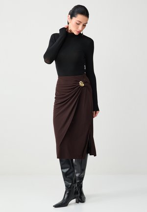 Kobieta w czarnym golfie, brązowej midi spódnicy z złotą broszką i czarnych wysokich botkach, stojąca na jasnym, jednolitym tle.