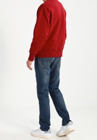 Sudadera de algodón roja con puños de canalé, combinada con jeans de mezclilla azules. Zapatillas blancas con detalles en azul claro. Vista trasera del conjunto.