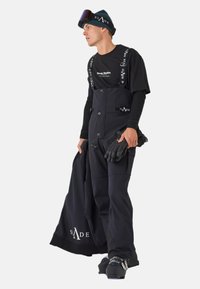 Schwarze Schneehosen mit verstellbaren Trägern, Seitentaschen und Knöpfen. Kombiniert mit einem schwarzen Langarmshirt und Handschuhen sowie Schuhwerk mit blauen Akzenten.