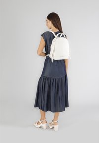 Zaino in pelle bianca con forma arrotondata e cerniere doppie, indossato sopra un vestito in denim scuro, che mette in risalto un design semplice e accenti minimalisti.