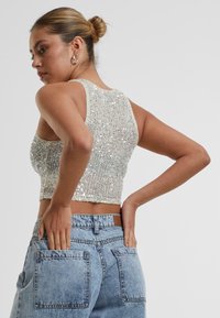 Jiskřivý stříbrný crop top s žebrovanou texturou, bez rukávů a přiléhavým střihem. Kombinován s světle modrými džíny s vysokým pasem.