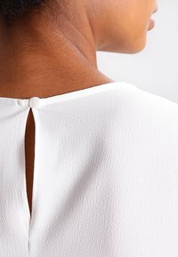 Haut blanc avec un tissu texturé, détail en clé de dos et une seule fermeture à bouton rond dans la nuque.