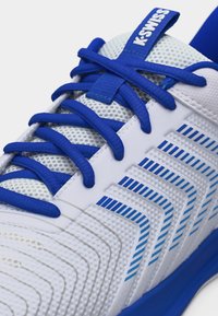 Κοντινό πλάνο λευκού αθλητικού παπουτσιού με μπλε κορδόνια, μπλε γεωμετρικές ρίγες στο πλάι και ετικέτα "K-SWISS" στο δίχτυ της γλώσσας.