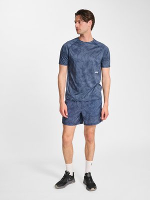 Ung mand med brunt hår iført matchende blå mønstret atletisk t-shirt og shorts, hvide ankelsokker og sorte løbesko, står.