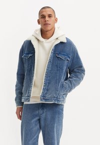 Denim jacket in faded blue with a white faux shearling collar, two chest pockets, and snap-button closure, worn over a cream hoodie.