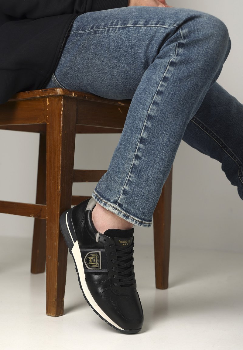 Schwarze Leder-Sneaker mit strukturierter Oberfläche, ausgestattet mit einer weißen Gummisohle und goldenen Logodetails. Getragen mit blauen Jeans.