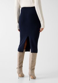 Jupe midi côtelée bleu marine avec une fente à l'arrière, associée à des bottes hautes taupe à talons aiguilles avec des accents dorés et une finition en daim texturé.