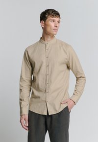 Lichtbeige overhemd met lange mouwen en een mandarin kraag, knoopsluiting aan de voorkant en subtiele textuur. Ggedragen met donkergrijze broek.