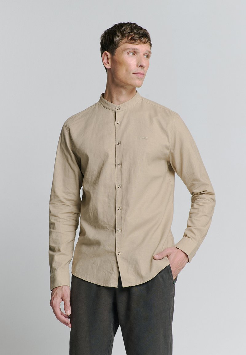 Lichtbeige overhemd met lange mouwen en een mandarin kraag, knoopsluiting aan de voorkant en subtiele textuur. Ggedragen met donkergrijze broek.