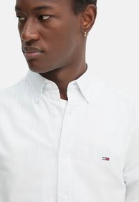 Camicia a righe azzurro chiaro con colletto, dotata di un piccolo logo ricamato sul lato sinistro del petto. Materiale in cotone, texture liscia.