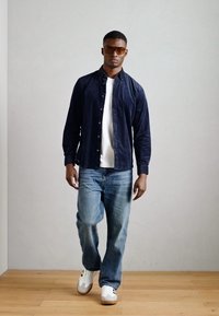 Mørk navy button-up skjorte over en hvid t-shirt, parret med lyseblå jeans og hvide sneakers. Modellen har brune, rektangulære solbriller på.