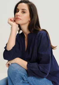 Granatowa lniana bluzka z bufkami i misternymi wycięciami wzdłuż dekoltu. Zestawiona z jasnoniebieskimi dżinsami.