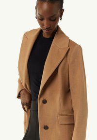 Camel-Wollmischungsmantel mit Reverskragen, doppeltknopfverschluss und aufgesetzten Taschen. Über einem schwarzen Oberteil getragen, glatte Textur.