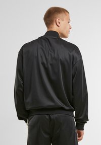 Veste zippée en satin noir avec col montant, manches longues et poignets côtelés. Texture lisse et coupe décontractée. Convient à diverses activités.