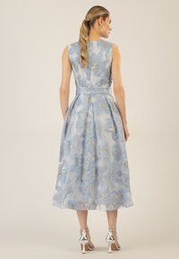 Ein ärmelloses knielanges Kleid aus hellblauem Stoff, das mit floralen Mustern und goldenen Akzenten versehen ist und eine gebundene Taille hat, wird mit silbernen High Heels kombiniert.