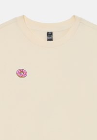 Cremfarbener T-Shirt mit Rundhalsausschnitt und kurzen Ärmeln, das auf der linken Brustseite eine pinkfarbene Donut-Stickerei hat.