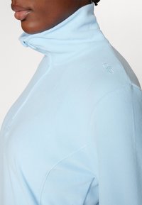 Hele sinine fliispull, millel on kõrge krae, pool-zip sulgemine ja diskreetne logo detail õlal. Pehme tekstuur.