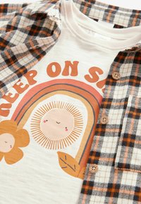 Kūdikių apranga, kurioje yra balta marškinėlių su saulės ir vaivorykštės grafiką, uždėta ant oranžinės ir juodos languotos flanelės marškinių su mygtukais.