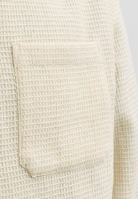 Gros plan d'un tissu en maille gaufrée couleur crème avec une poche patch rectangulaire cousue sur sa surface.