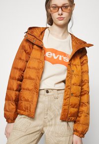 Padded oranžová bunda s kapucňou, s prešívaným dizajnom a elastickými manžetami. Nosená cez béžové tričko a svetlé vzorované nohavice.