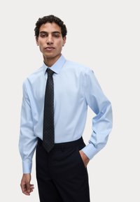 Jasnoniebieska koszula z długim rękawem i klasycznym kołnierzykiem, połączona z czarnym krawatem w małe zielone kropki oraz ciemnymi spodniami.