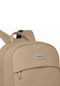 Sac à dos en tissu beige avec une texture lisse, comprenant une poche avant, une poignée supérieure et le logo de la marque sur un patch argenté.
