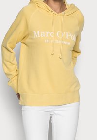 Persona che indossa un hoodie giallo di Marc O’Polo con lacci e pantaloni bianchi, in piedi contro uno sfondo chiaro e uniforme.
