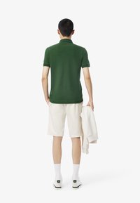 Polo verde a maniche corte, pantaloni corti color crema, calze bianche e scarpe da ginnastica. Un sweatshirts beige è appoggiato sul braccio.