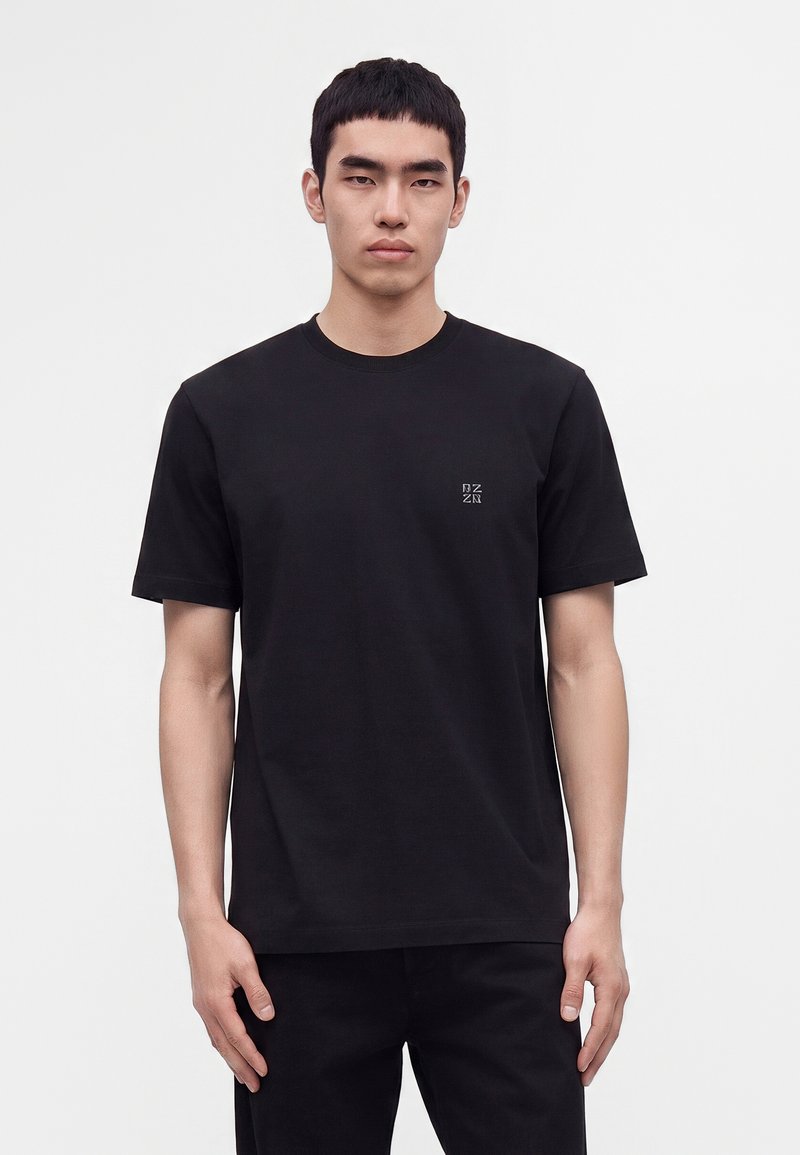 Jeune homme portant un t-shirt noir uni à manches courtes avec un petit logo blanc sur le côté gauche de la poitrine, se tenant devant un fond blanc.