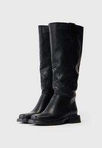 Um par de botas pretas de couro até ao joelho com saltos baixos em um fundo claro e liso.
