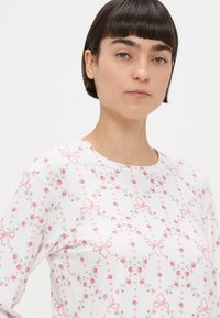 Λευκό μπλουζάκι με μακριά μανίκια, διακοσμημένο με ροζ λουλούδια και φιόγκους, κατασκευασμένο από απαλό, υφασμάτινο ύφασμα με υφή. Στρογγυλή λαιμόκοψη και άνετη εφαρμογή.