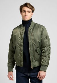 Giacca bomber verde oliva con zip frontale, polsini a costine e molteplici tasche; indossata sopra un maglione a collo alto in maglia blu navy.