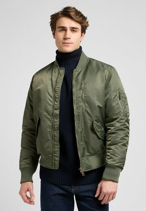 Olive green bomber jacket with a front zip, ribbed cuffs, and multiple pockets; worn over a navy knitted turtleneck sweater.