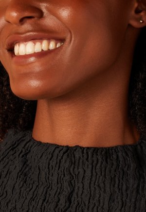 Top en tissu noir texturé avec un design froissé, porté par une personne souriante. Présente une peau lisse et une petite boucle d'oreille à l'oreille.