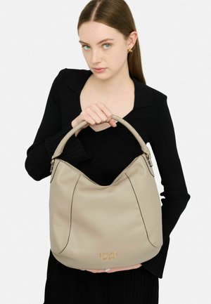 Borsa hobo in pelle beige con logo dorato, cuciture a contrasto e una maniglia morbida. Il modello tiene la borsa, mettendo in evidenza la sua forma morbida e il design.